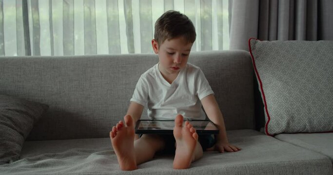 Curious cute preschool kid boy using digital tablet technology device lying on carpet floor alone. Small child hold pad computer surfing internet play game at home. Children tech addiction concept