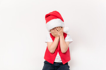 Portrait of crying little child in red Santa Claus hat isolated on white background. European 5 year boy. Without gifts. Face is covered with hands. Bad mood. Christmas. Close-up. Sleepy or tired kid