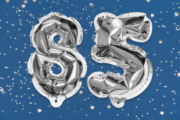 Silver foil balloon number, digit eighty five on a blue background with sequins. Birthday greeting...