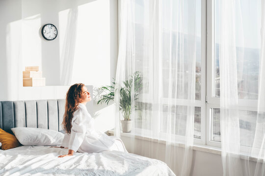  Woman in white bathrobe sitting near the big white window on bed after waking up.