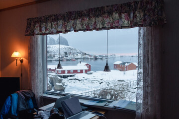 Interior of lamp shining with view fishing village on winter through window and laptop on desk in scandinavian house