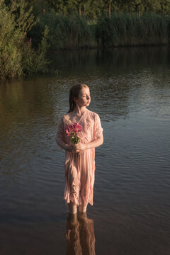 Girl in pink dress holding flowers standing in water in lake in evening light
