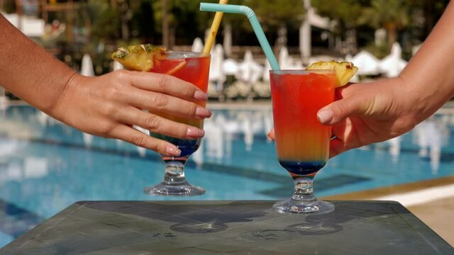 Close-up of two young women taking glasses with colorful cocktails, tubes and pineapple slices on the background of the pool. Summer vacation on the beach.