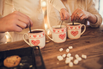 Fototapeta na wymiar Family: mom, dad and daughter in white sweaters cook and drink cocoa with marshmallows. Closeup hands and cups. Christmas concept
