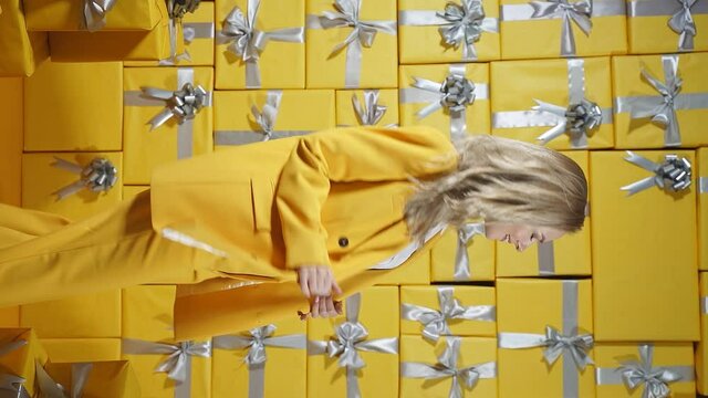 A pretty blonde is impressed by the photo zone from gift boxes. An emotional woman is happy to pose in front of the camera on a yellow background.