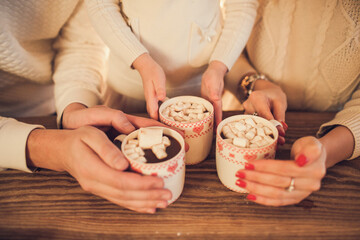 Obraz na płótnie Canvas Family: mom, dad and daughter in white sweaters cook and drink cocoa with marshmallows. Closeup hands and cups. Christmas concept