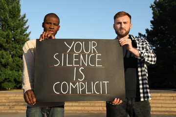 Men holding sign with phrase Your Silence Is Complicit outdoors. Racism concept