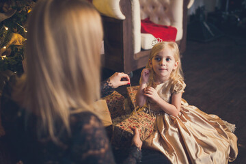 A little girl in an elegant dress and her mom open a gift box under the Christmas tree