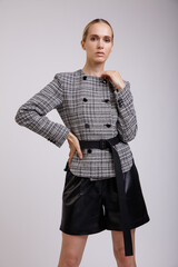 High fashion photo of a beautiful elegant young woman in a pretty grey jacket, black leather shorts, belt posing on white background. Studio Shot. Slim figure.