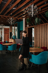 a business woman in a black dress is standing in a cafe with a laptop and working