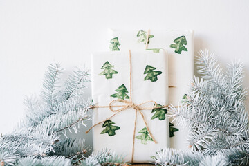 the New Year's gift is packed in festive paper with watercolor Christmas trees on a wooden table against a white wall with white spruce branches
