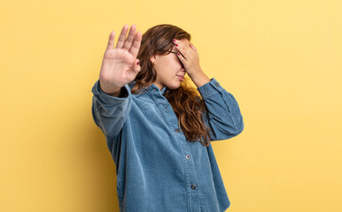 hispanic pretty woman covering face with hand and putting other hand up front to stop camera,...