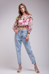 High fashion photo of a beautiful elegant young woman in a pretty blouse top with floral pattern in red, blue jeans posing over white, soft gray background. Slim figure. Studio Shot.