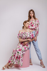 Two high fashion models in long dress with a red floral pattern, blouse, blue jeans. Beautiful young women. Studio shot. White background. Make up, hairstyle. Model is sitting on a pink cube cylinder