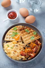 Waffles served with scrambled eggs and tomatoes, vertical shot over light-blue stone background, elevated view