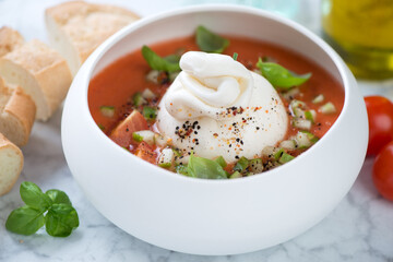 Tomato gazpacho soup served in a white bowl with burrata cheese, green basil and baguette, middle close-up, selective focus