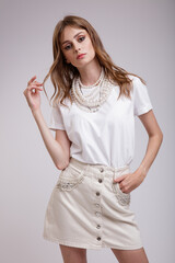 High fashion photo of a beautiful elegant young woman in a pretty white denim skirt, t-shirt, pearl necklace posing over soft gray background. Slim figure. Studio shot