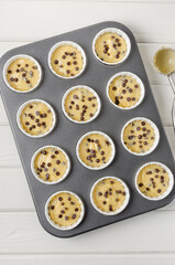 Obraz premium Raw dough for muffins with chocolate chips in baking form on a white wooden background. Recipe step by step. Top view.