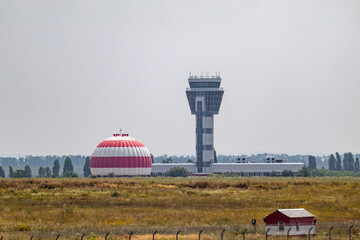 International airport control tower and radome or aviation radar dome building in red and white stripes in grass field with gray sky distant view in summer - obrazy, fototapety, plakaty