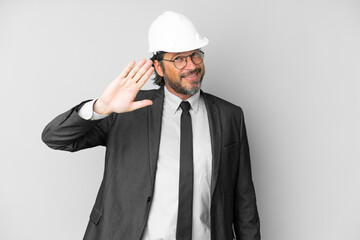 Young architect man with helmet over isolated background saluting with hand with happy expression