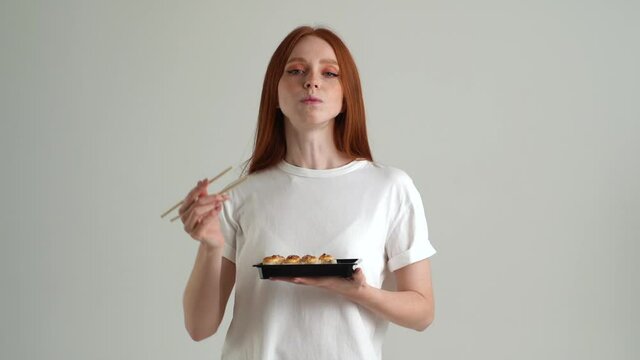 Portrait of cheerful pretty young woman eating sushi rolls with chopsticks and looking at camera standing on white isolated background in studio. Happy Caucasian redhead female eats Asian food.