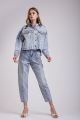 High fashion photo of a beautiful elegant young woman in a pretty denim jeans oversize jacket with rhinestones, pants posing over white, soft gray background. Slim figure.  Studio Shot.