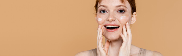 Excited freckled woman with cosmetic cream on cheeks looking at camera isolated on beige, banner