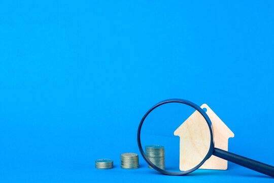 Wooden house keychain, magnifier and stacks of coins on a blue background. Home search and buying concept