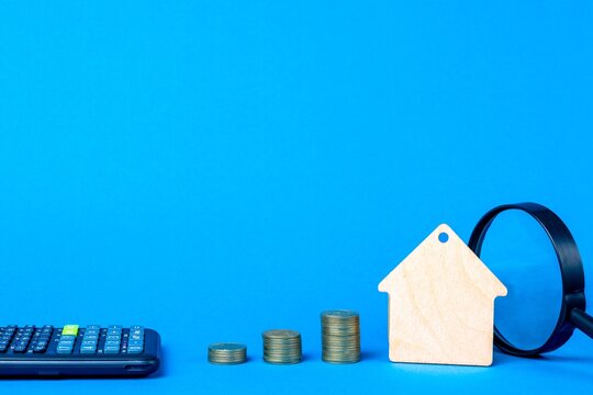 Wooden house keychain, stacks of coins, calculator and magnifier on a blue background. Home search and buying concept
