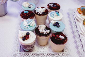 tiramisu sprinkled with cocoa and garnished with blueberries, currants, caramelized snowflakes, meringues and marshmallows, in a clear plastic glass on a tray