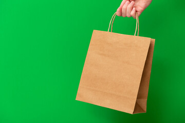 Female hand holding brown craft blank shopping bag isolated on green background. Black friday sale,...
