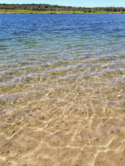 Landscape of lake and forest in the far and texture of crystal clear light Blue, green and turquoise transparent water with fine white sand base with bright reflection of sun