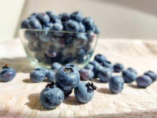 Big, ripe cultivated blueberries or highbush blueberries in a glass bowl and on table covered with gray tablecloth with white background in bright sunlight