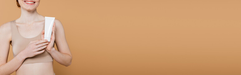 Cropped view of positive freckled woman holding tube with lotion isolated on beige, banner