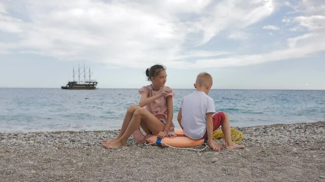 A boy and a girl of 8 years old are sitting on the seashore on a lifebuoy, against the background of a ship floating in the sea.