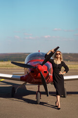 Fashionable women in beautiful classy black dress and beautiful sunglasses posing on a red private plane and blue sky in background