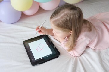 A little girl draws with a stylus on a tablet. A little girl uses a tablet  at home against the background of balloons.Digital girl