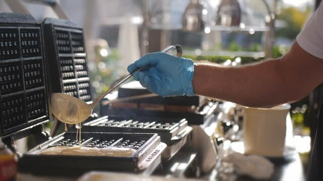 Close-up of a chef in gloves pouring dough on a waffle iron. Cooking waffles outdoors in a street restaurant.