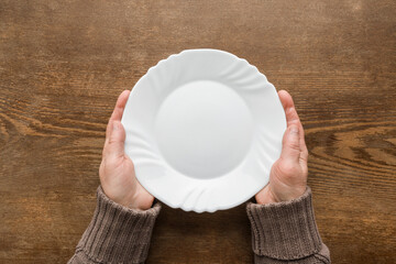 Mature woman hands holding white empty plate on dark brown wooden table background. Closeup. Point of view shot. Meal waiting concept. Top down view. - Powered by Adobe