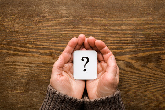 White card of question mark on mature woman palms on dark brown wooden table background. Point of view shot. Concept of what to do in old ages. Pensioner issues. Closeup. Top down view.