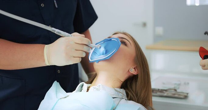 A dentist wearing gloves drills a tooth to a female patient with a cofferdam system in a modern dental office. The assistant puts a light seal to the patient. Dental treatment. High-quality 4k video. 