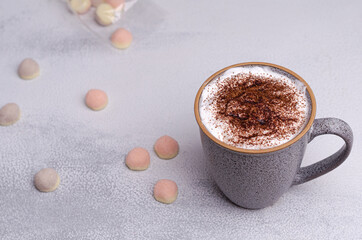 A gray coffee mug with cream on a light gray table with small candies. Delicious breakfast and time to relax.