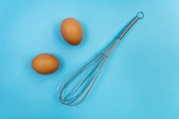 Two brown eggs and a whisk for whipping on a blue background. Natural products and kitchen 

equipment