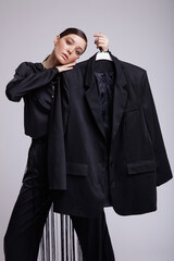 High fashion photo of a beautiful elegant young woman in a pretty black blouse, fringe trousers, accessories over gray background. Studio Shot. Holds  jacket in hands on a hanger