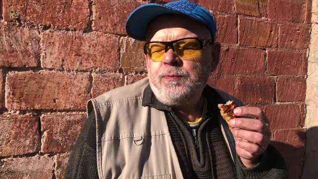 Caucasian senior man eating chopped meat in lavash while sitting against red brick wall at sunny day