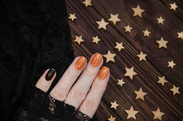 Female hand with orange glitter nails holds orange nail polish on the dark starry background.