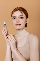 Portrait of freckled woman holding cosmetic serum isolated on beige