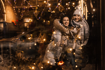 two beautiful young women smiling and hugging, reflected in a showcase with Christmas lights.