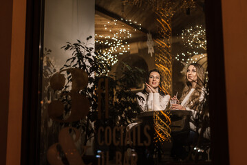 two beautiful young women are sitting at a table in a cafe and drinking coffee. view through the window and beautiful blurred lights