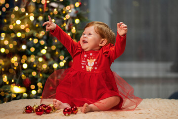 A little girl with red hair in a red dress sits and laughs against the background of a Christmas tree. The inscription on the clothes: Happy New Year.